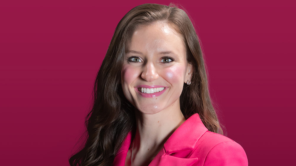 Megi Maci wearing a magenta suit and smiling while standing in front of a maroon